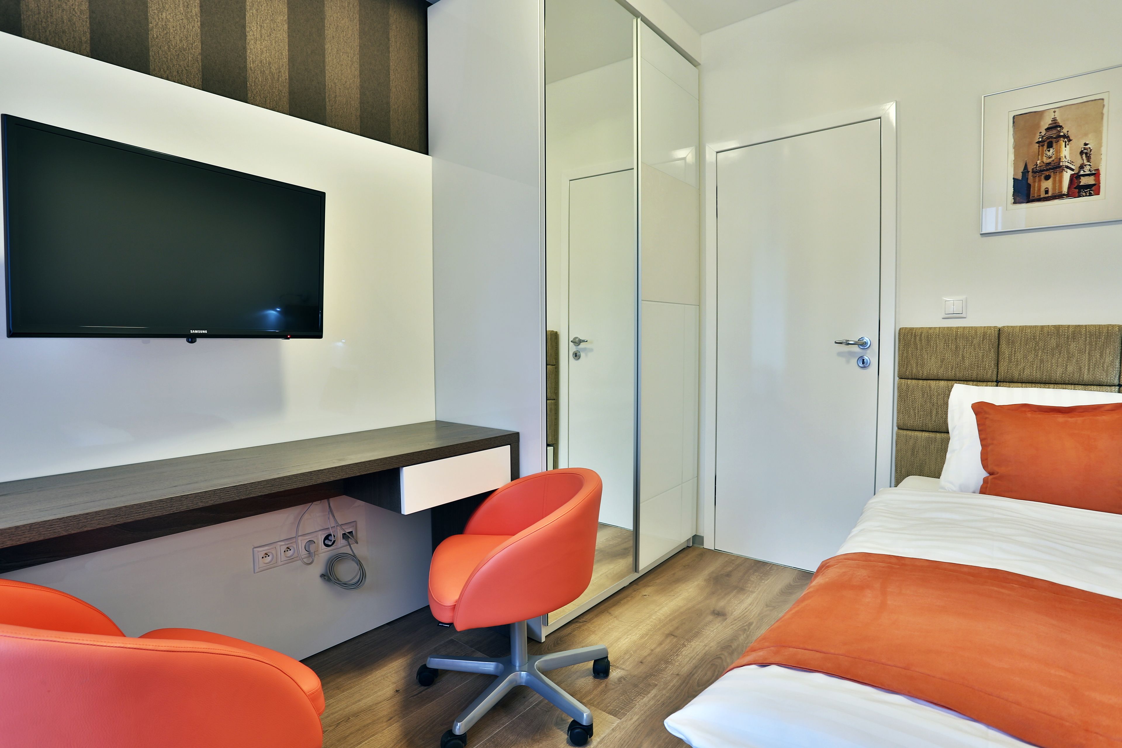 A modern hotel room with a bed, a desk with a chair, a TV, and a wardrobe.
