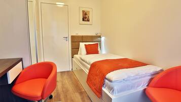 Quarto com uma cama, duas cadeiras laranja, uma escrivaninha de madeira e uma porta.