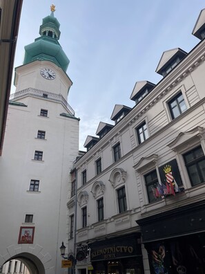A building with a clock tower, a sign for 'KLENOTNÍCTVO BENIX', and flags.