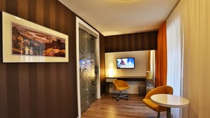 Una habitación de hotel con suelo de madera, papel tapiz a rayas, un cuadro enmarcado, un televisor y sillas naranjas.