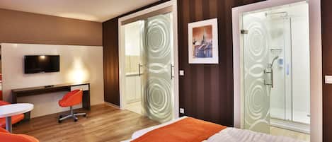 A hotel room with a bed, a desk with a chair, a TV, and a shower area.