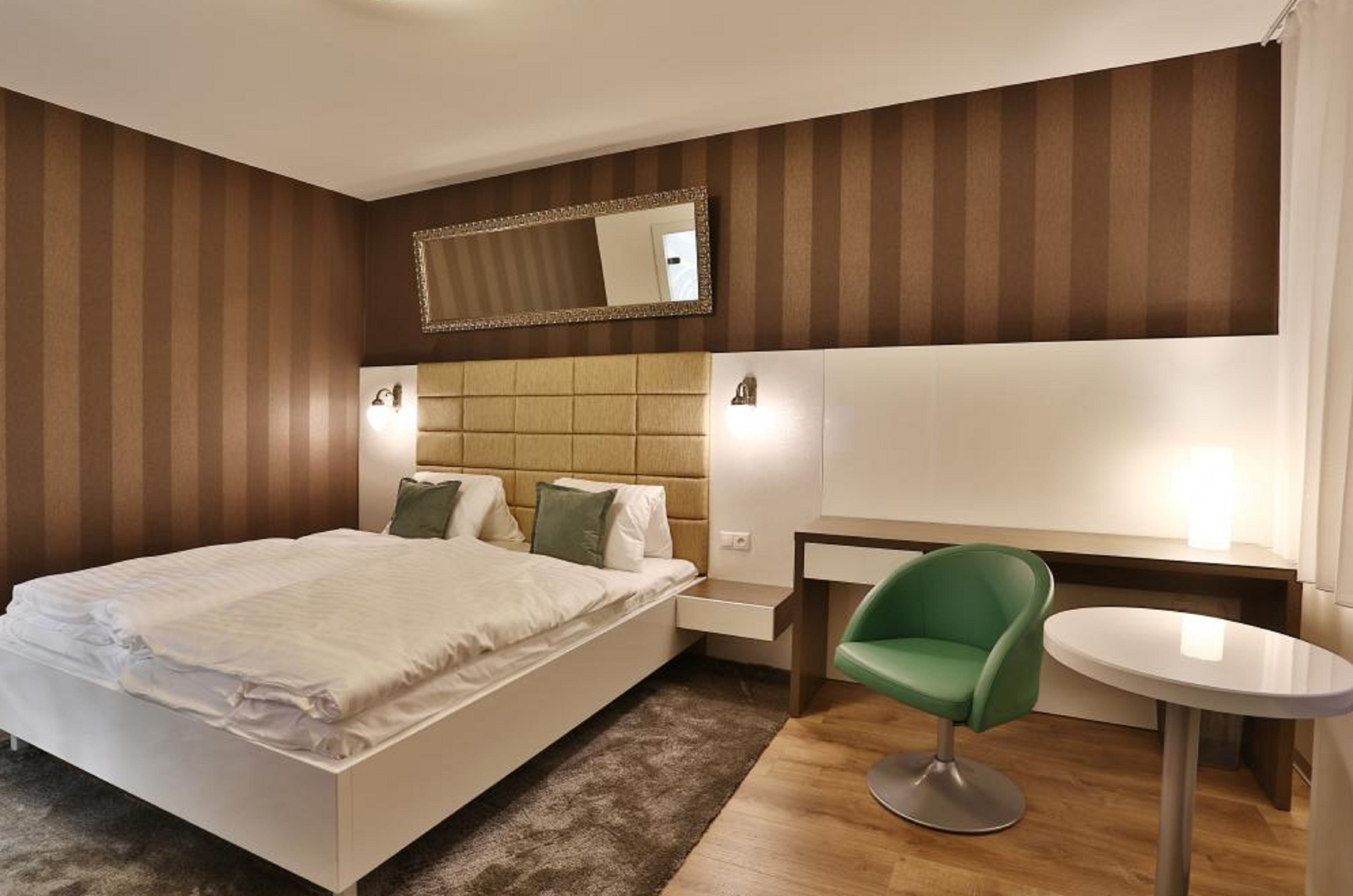 Quarto de hotel moderno com uma cama grande, uma cadeira verde, uma mesa pequena e um espelho na parede.