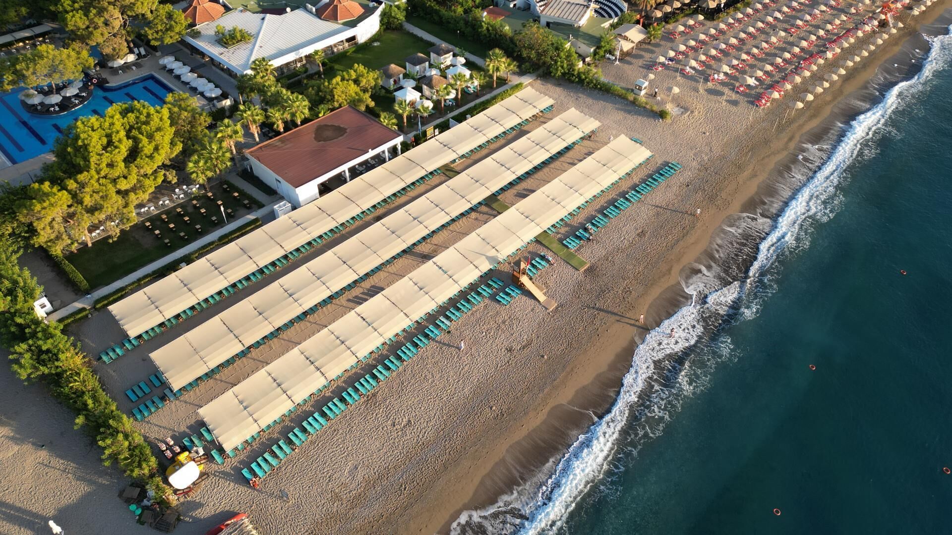 Vista aèria d'un complex turístic a la platja, amb piscina, parasols i gandules.