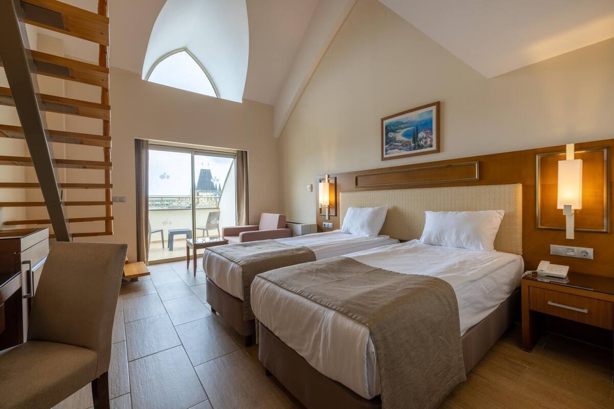 A hotel room with two beds, a desk, a chair, a desk lamp, a painting, and a balcony with a view.