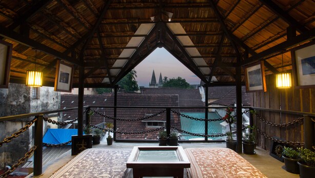 A covered balcony with a view of a cityscape, featuring a wooden ceiling, chain-link railing, and potted plants. - Niyati - Boutique Stay (Kochi)