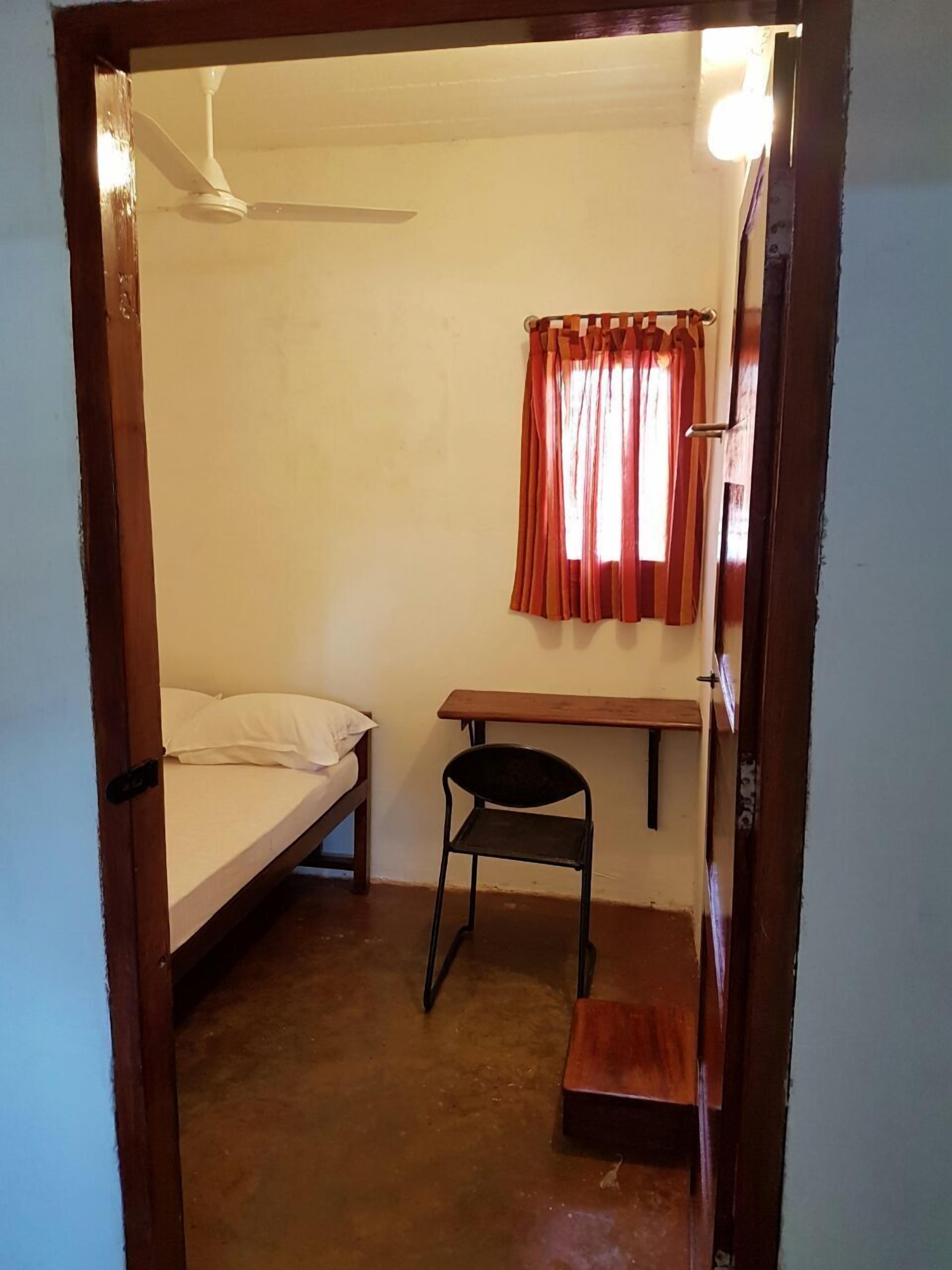 Eine kleine Einzelunterkunft mit Bett, Stuhl, Schreibtisch und einem fenster mit Vorhängen.