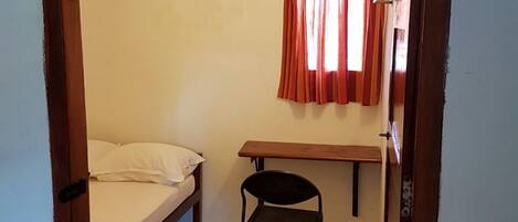 Eine kleine Einzelunterkunft mit Bett, Stuhl, Schreibtisch und einem fenster mit Vorhängen.