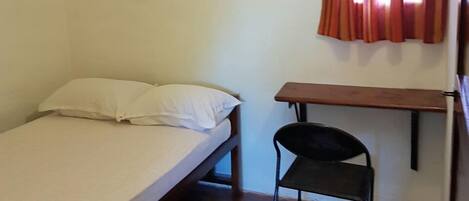 Una habitación pequeña con cama individual, ventilador de techo, ventana con cortinas rojas, un escritorio de madera y una silla negra.