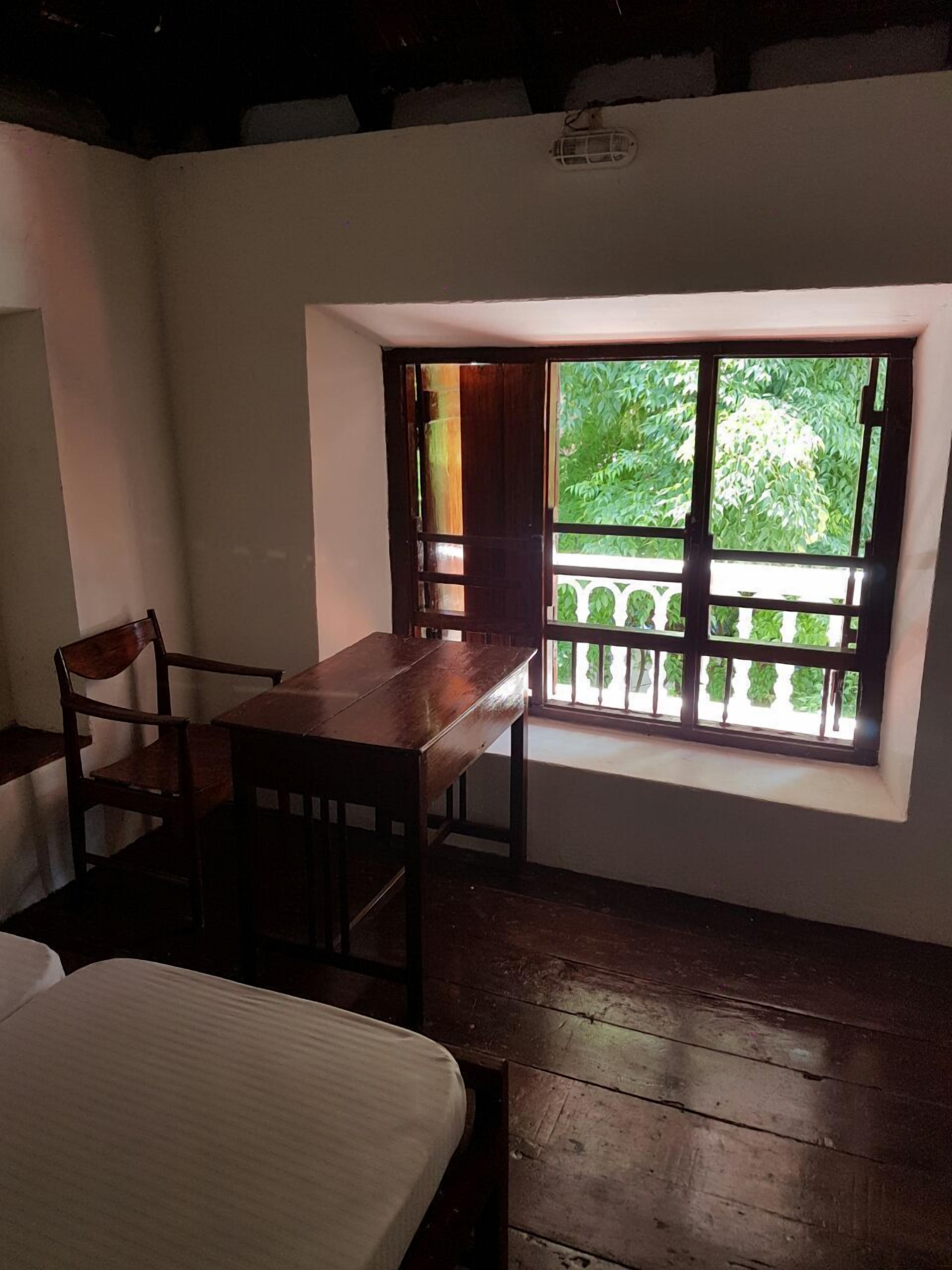 A small room with a wooden table and chairs, a window with a view of greenery, and a white bed.