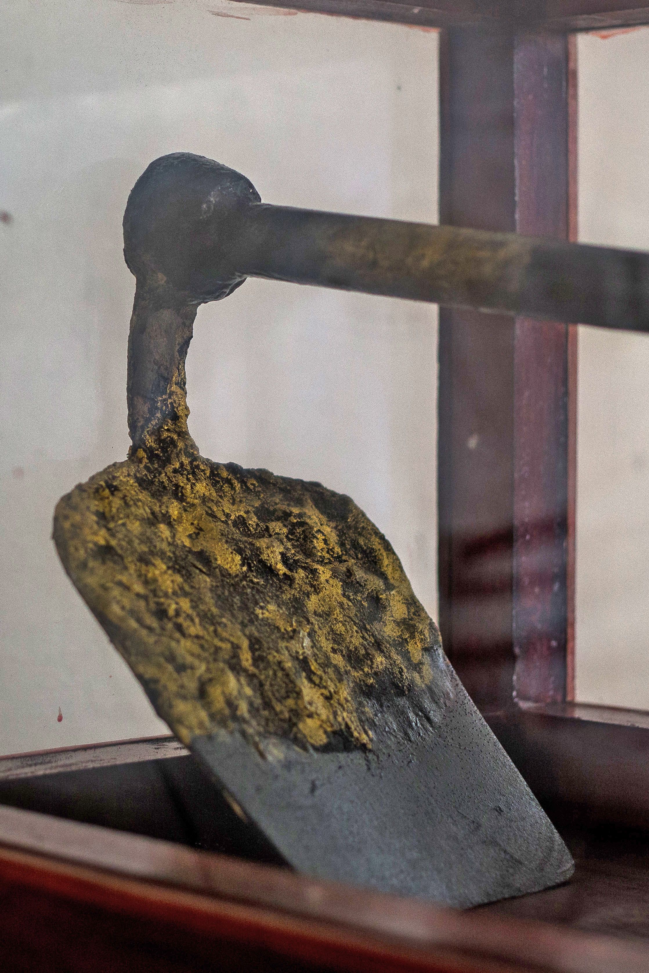 A metal object with a curved handle and a flat base, possibly a tool or utensil, displayed in a glass case.