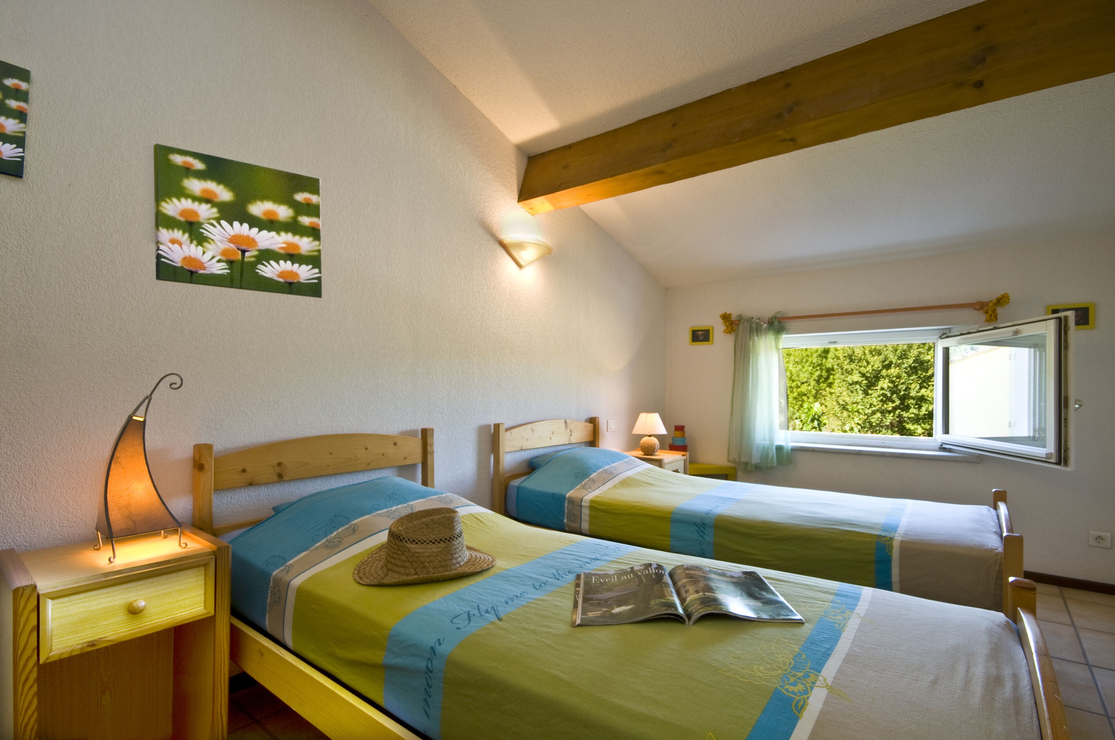 A room with two beds, a nightstand with a lamp, a window with a view of greenery, and a wall-mounted picture of daisies.