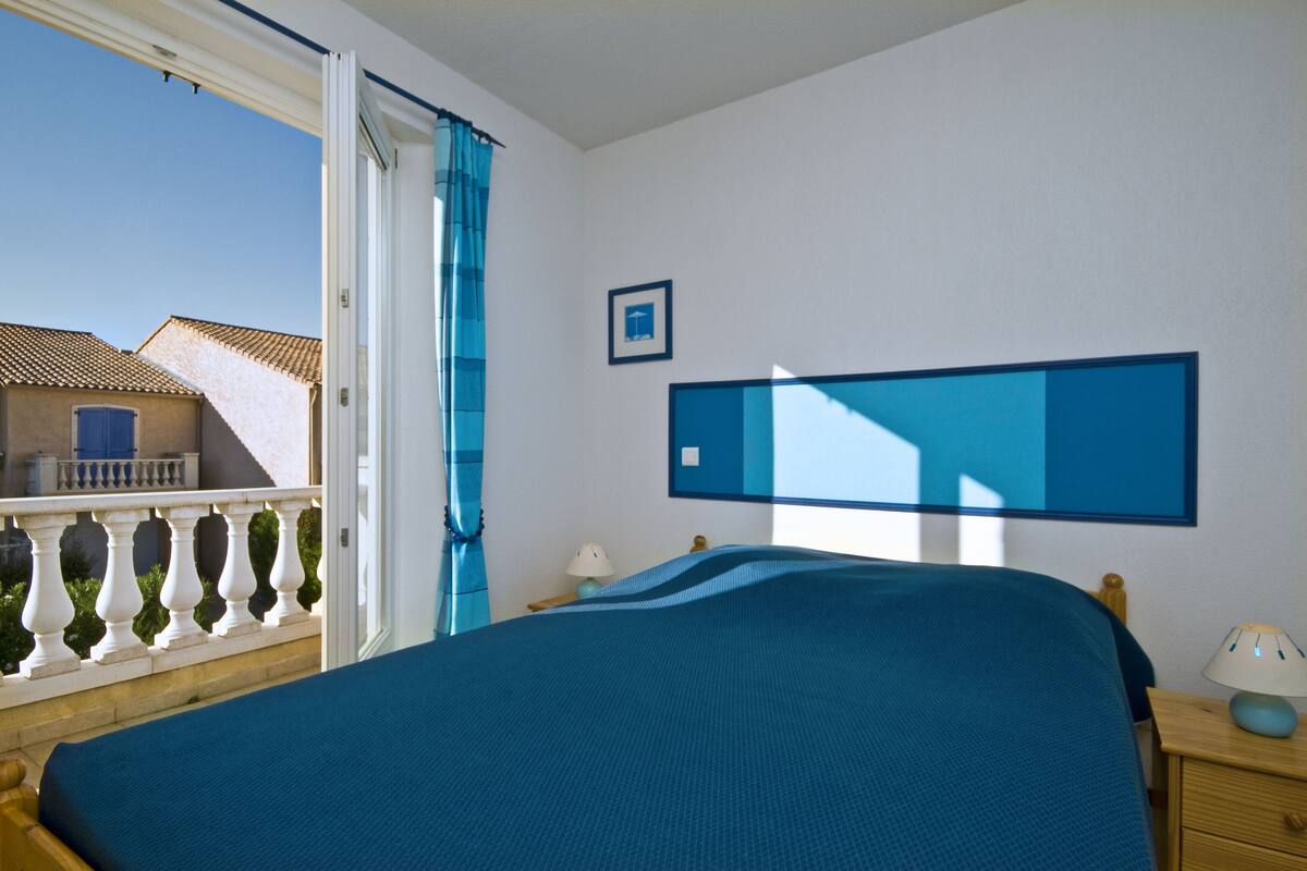 A bedroom with a bed, a nightstand, a lamp, a picture frame, and a view of a building outside.