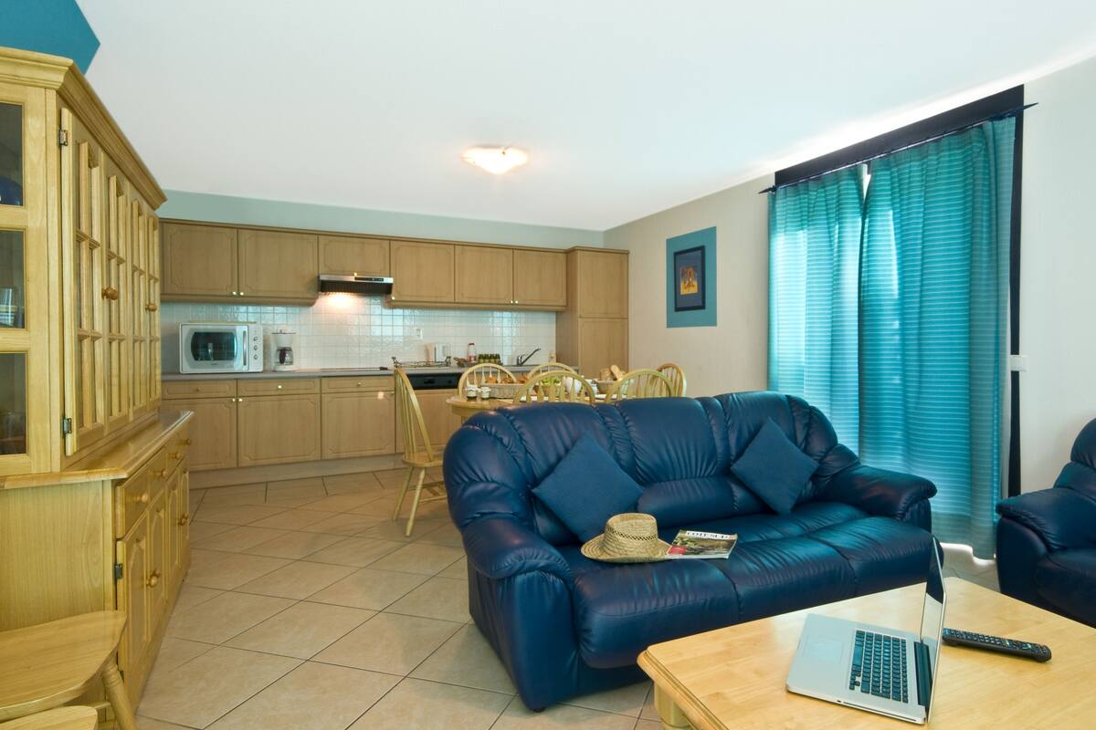 A living room with a blue sofa, a wooden coffee table, and a laptop. There is a kitchen in the background with wooden cabinets and a microwave.