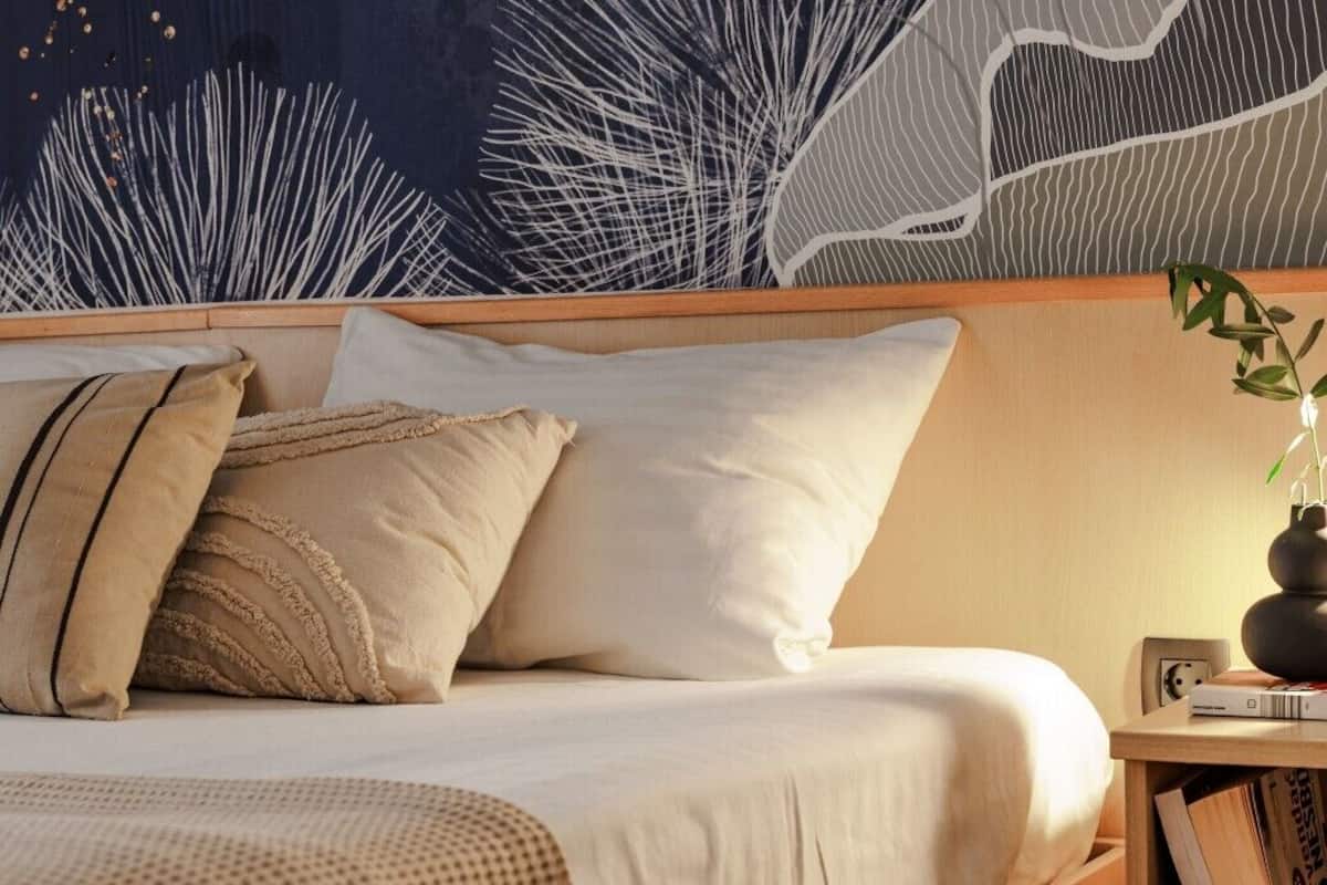 Ein ordentlich bezogenes Bett mit weißer und beiger Bettwäsche, ein hölzerner Kopfteil und eine Wand mit Blättermotiv.