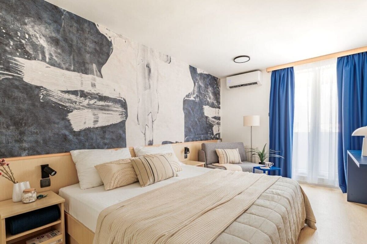 Ein modernes Schlafzimmer mit einem großen Bett, einem Nachttisch, einer Lampe und Blick ins Freie durch ein Fenster mit blauen Vorhängen.