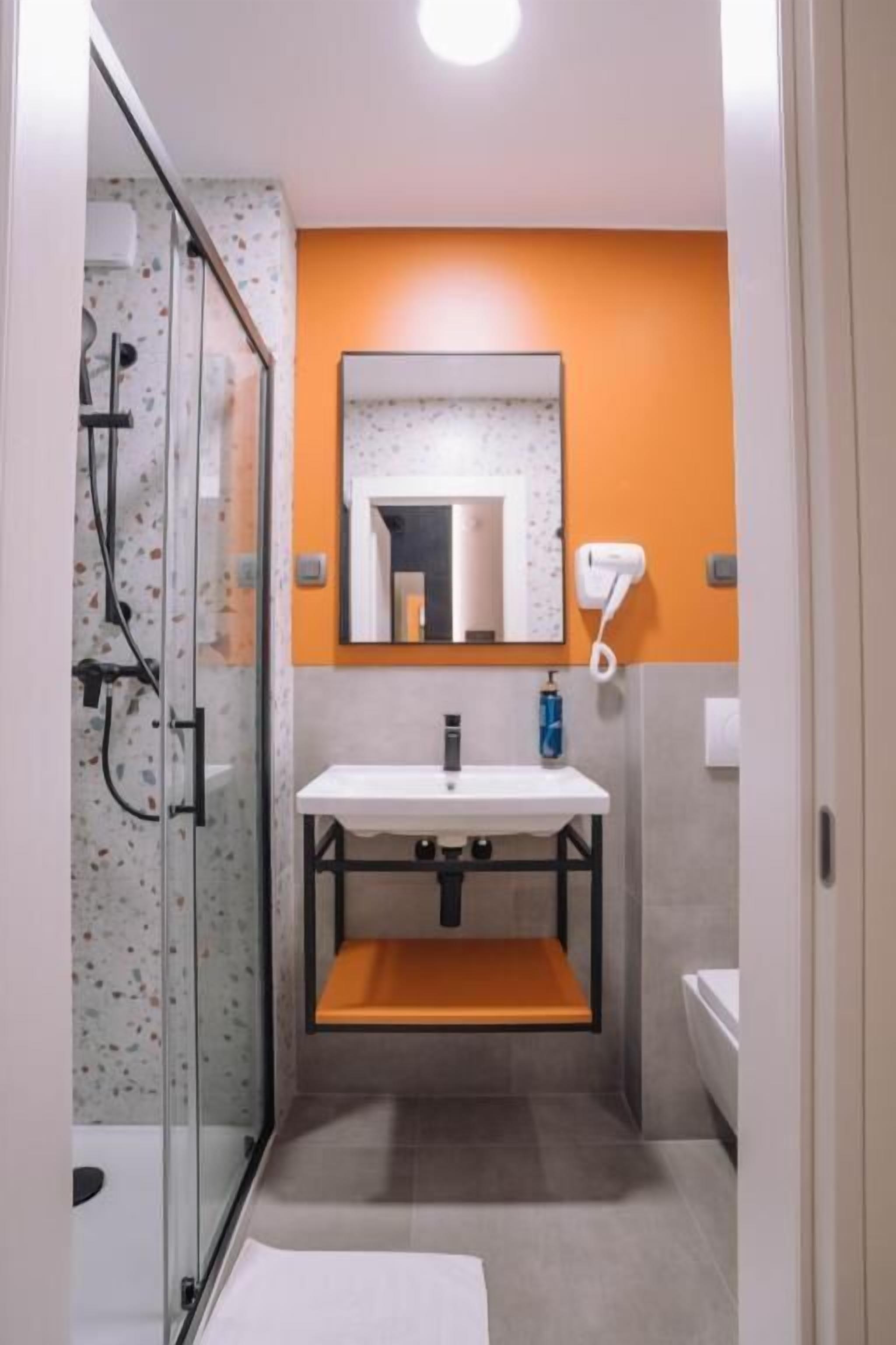 Ein modernes Badezimmer mit Duschkabine aus Glas, einem weißen Waschbecken mit Ablagefläche in Orange und einem Spiegel über dem Waschbecken.