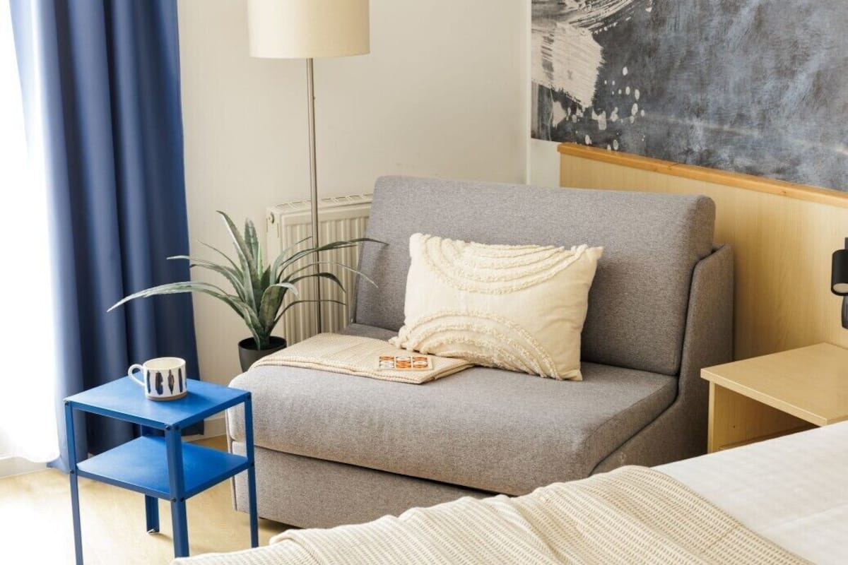 Ein Zimmer mit einem grauen Sofa, einem kleinen blauen Beistelltisch mit einer Tasse, einem weißen Kissen, einer Lampe und einem Bild an der Wand.
