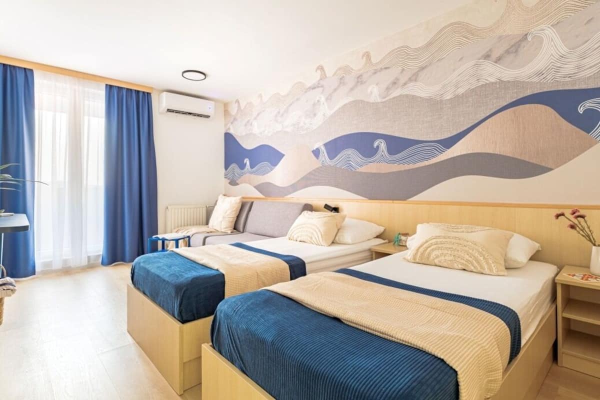 Ein Hotelzimmer mit zwei Betten, einem großen, abstrakten Wandbild, einem Fenster mit blauen Vorhängen und einem Nachttisch mit Blumen.
