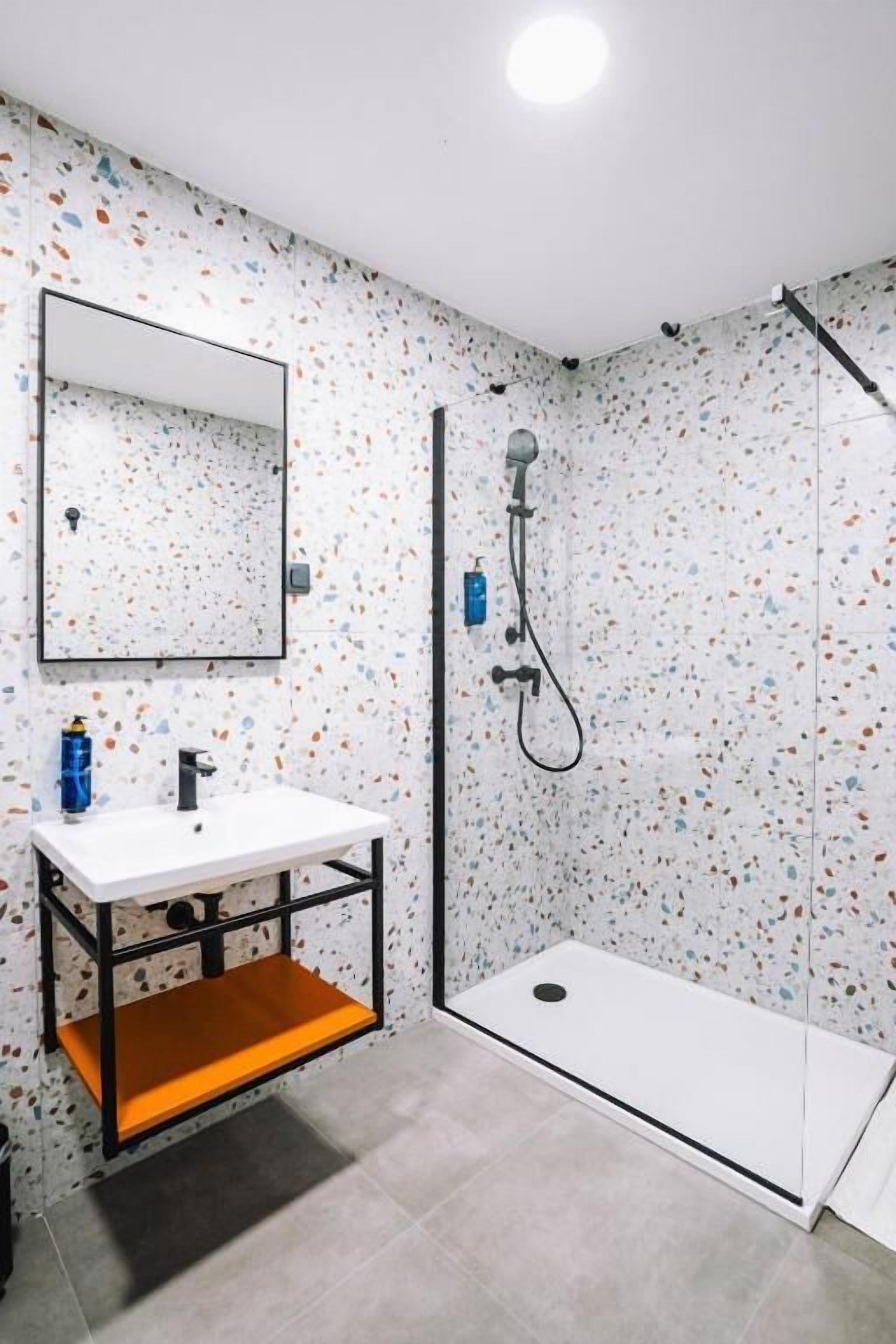 Un bagno con un lavandino bianco, uno specchio, una doccia con porta in vetro e un pavimento in terrazzo.