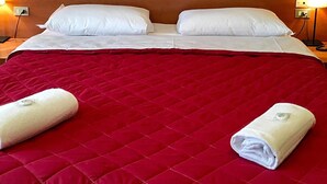Una habitación de hotel con una cama grande, dos almohadas blancas, una colcha acolchada roja y un cuadro de ángeles en la pared.