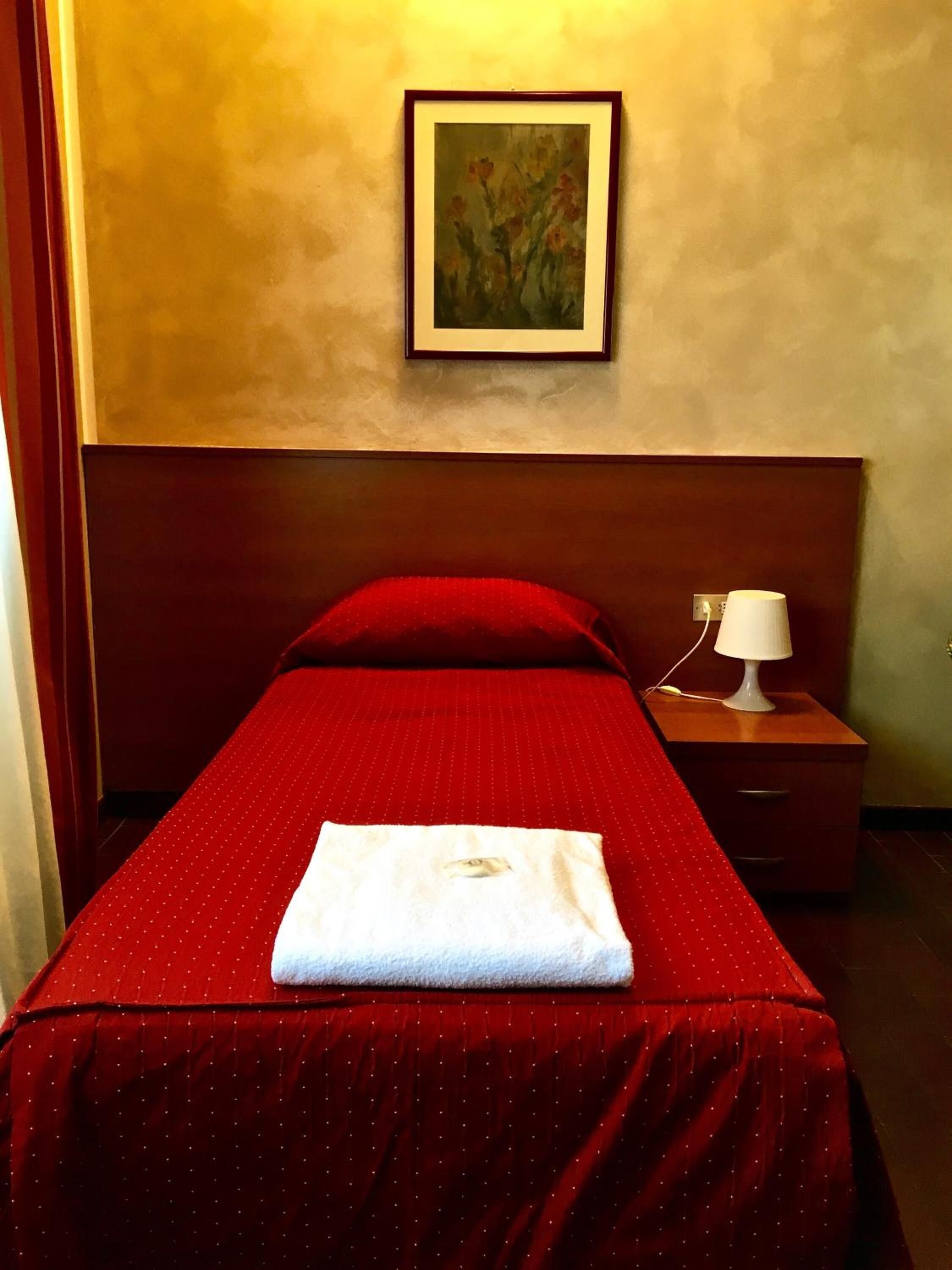 Une chambre d’hôtel avec un lit simple, une couvre-lit rouge, une serviette blanche, une table de chevet avec une lampe et un tableau au mur.
