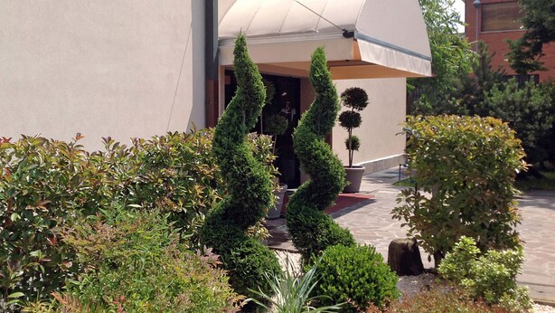Un jardín ajardinado con setos recortados y un árbol en maceta frente a un edificio con techo curvo.