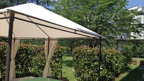 Un espacio al aire libre con techo, mesas y sillas, rodeado de follaje y un edificio al fondo.