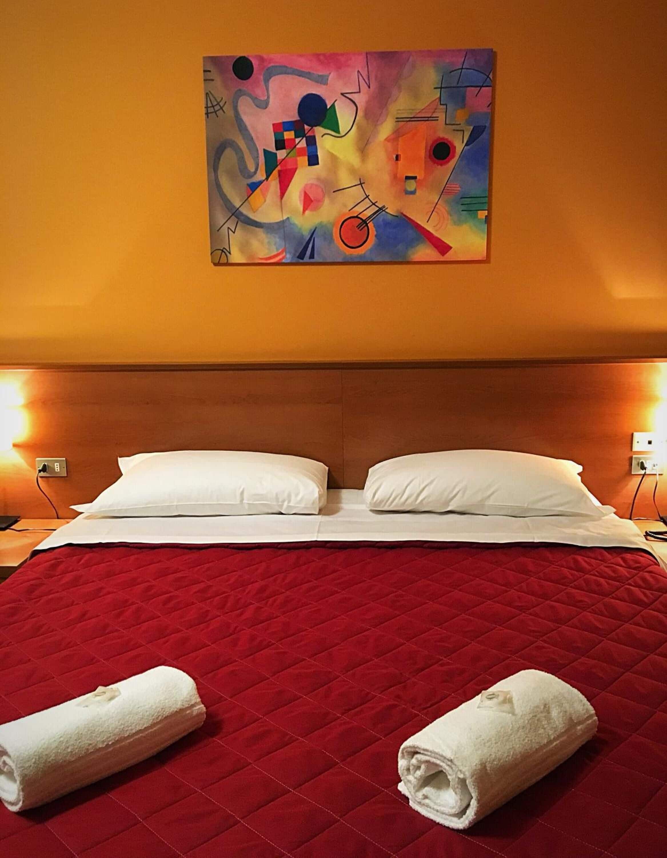 Ein Hotelzimmer mit einem großen Bett, einer roten gesteppten Bettdecke, zwei weißen Kissen und einem farbenfrohen abstrakten Gemälde an der Wand.