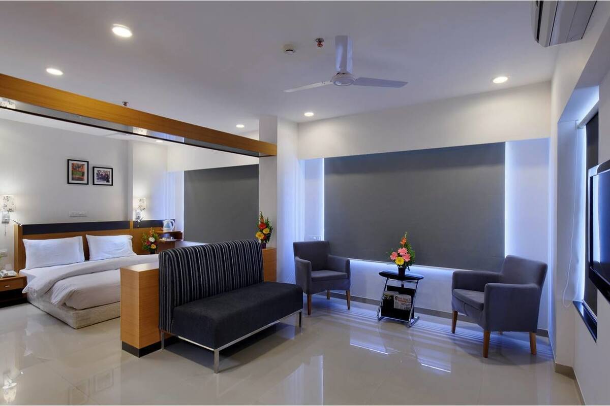 A modern hotel room with a bed, a sofa, two armchairs, a small table with flowers, and a large window.