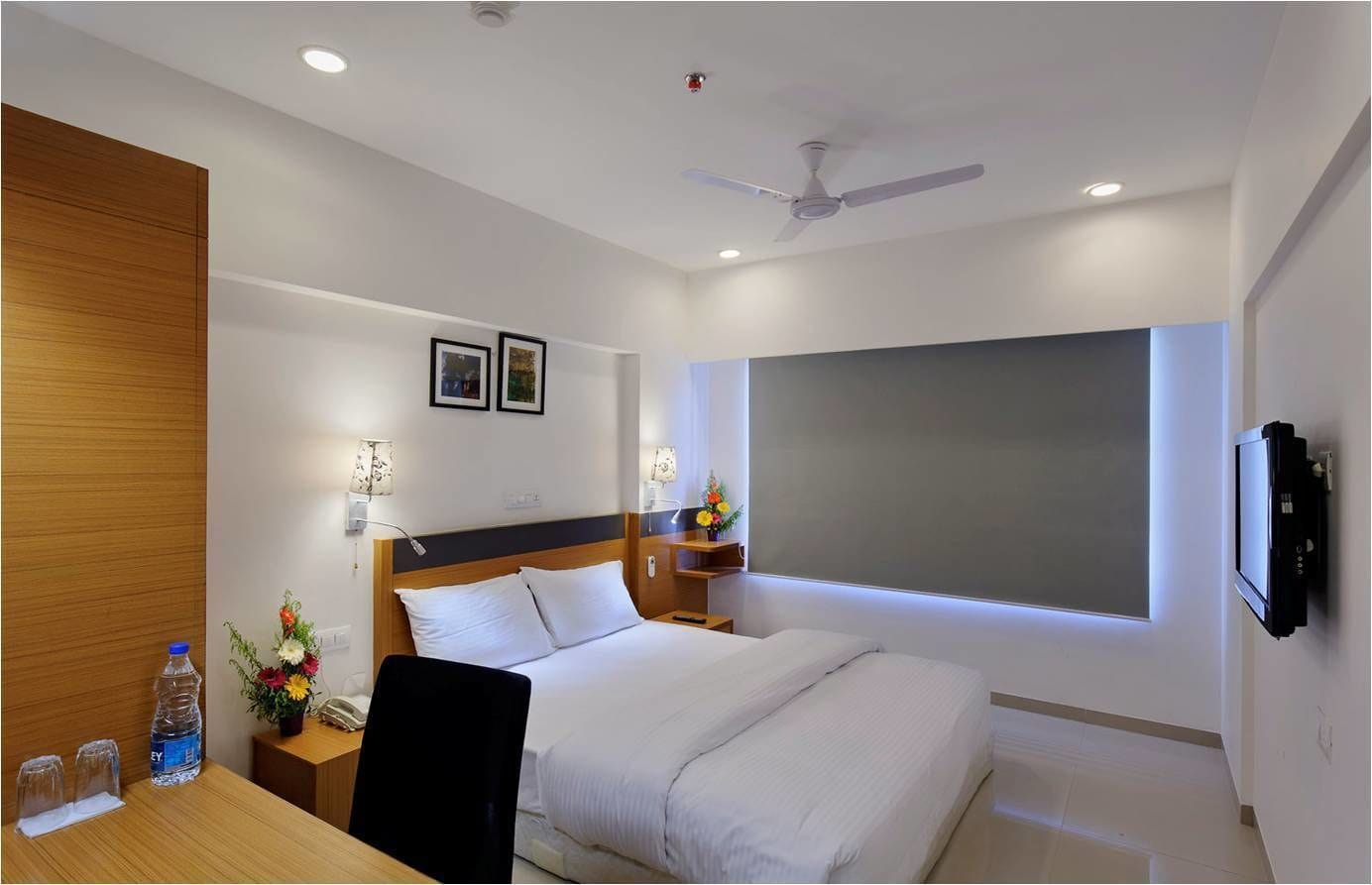 Quarto de hotel com cama, mesas de cabeceira, escrivaninha, cadeira, ventilador de teto, televisão e uma janela grande.