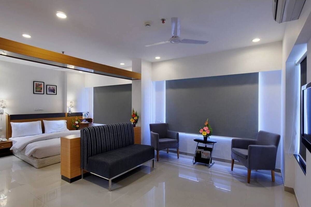 A modern hotel room with a bed, a sofa, two armchairs, a small table with a vase of flowers, and a large window.