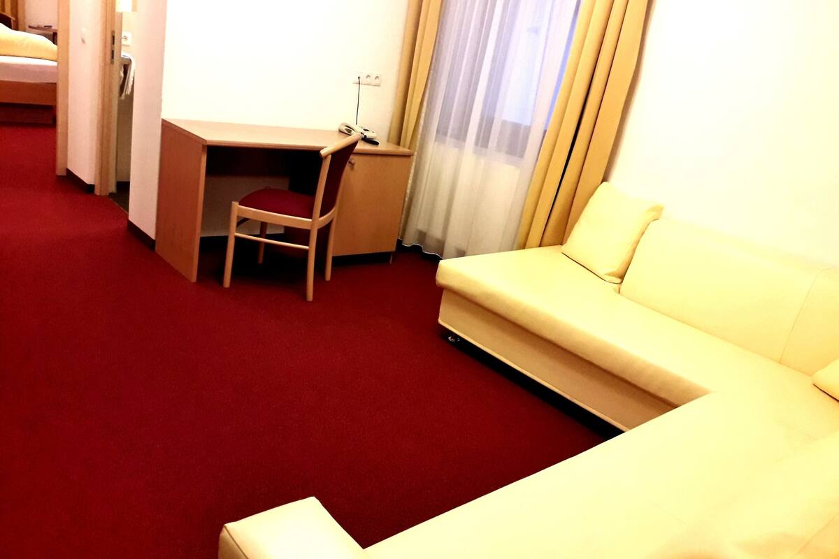 A hotel room with a red carpet, a white sofa, a desk with a chair, and a bed in the background.
