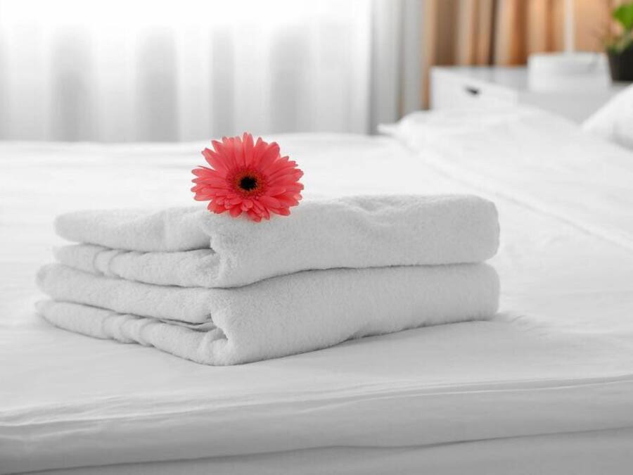 A neatly made bed with folded white towels and a single red flower on top.