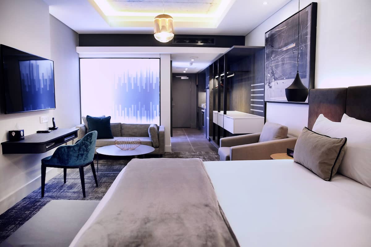 A modern hotel room with a large bed, a desk, a chair, and a sofa. There is a large window with a view of the city.
