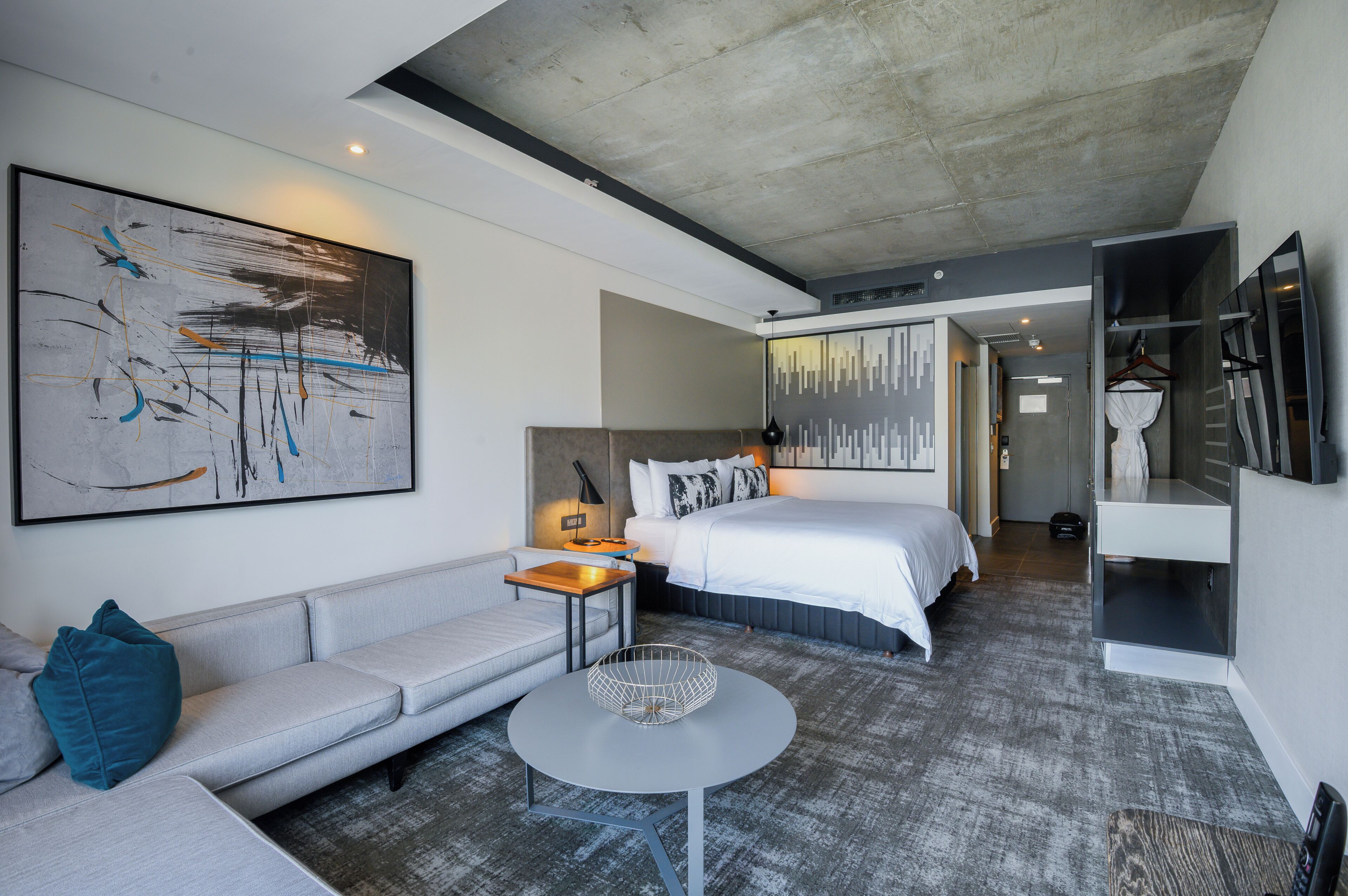 A modern hotel room with a large abstract painting, a bed with white bedding, a grey sofa, a small round table, and a grey carpet.