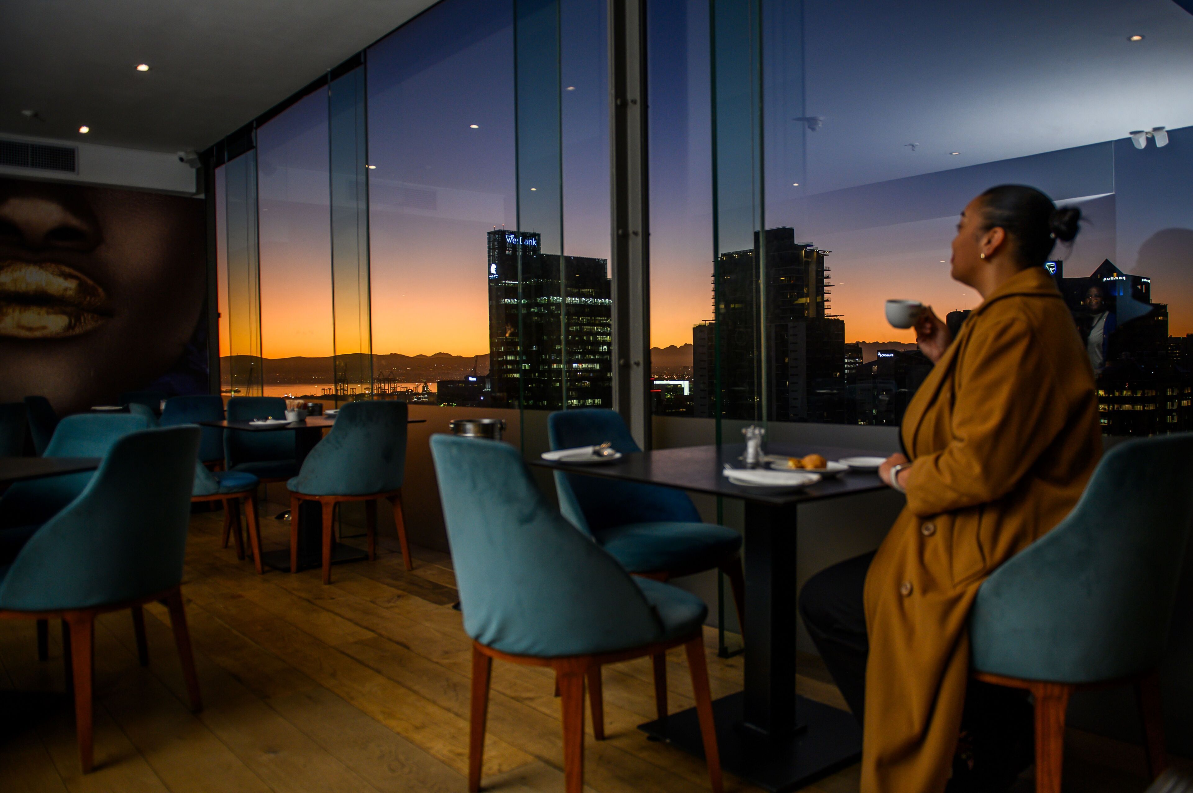 A modern dining area with a city view at sunset, featuring teal chairs, a wooden table, and a large window.