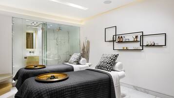 A modern spa room with two beds, a glass shower area, and shelves with toiletries.