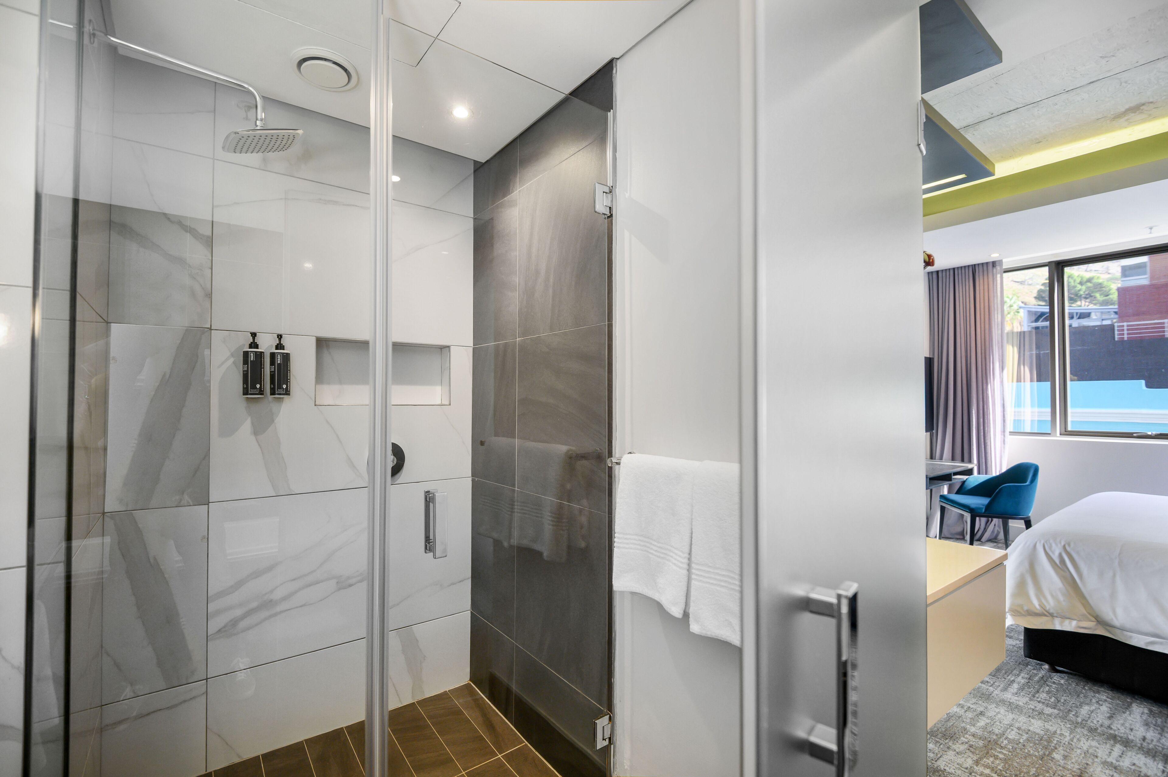 Ein modernes Hotelzimmer mit einer gläsernen Duschkabine, einem Bett mit weißer Bettwäsche, einem Schreibtisch mit Stuhl und Blick ins Freie durch ein großes Fenster.