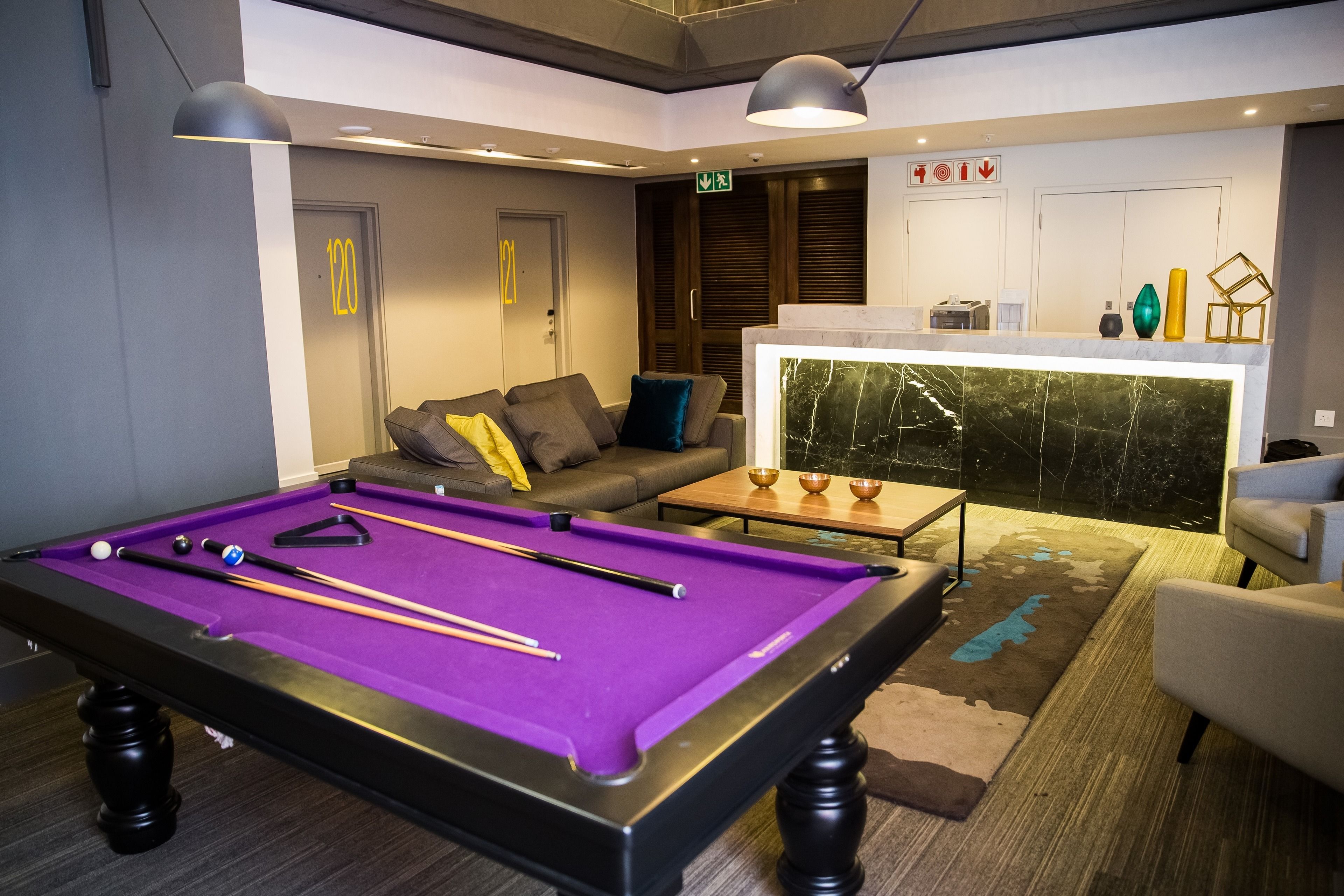 A pool table with a purple felt surface, a sofa with cushions, a coffee table, and a bar area with marble backsplash.