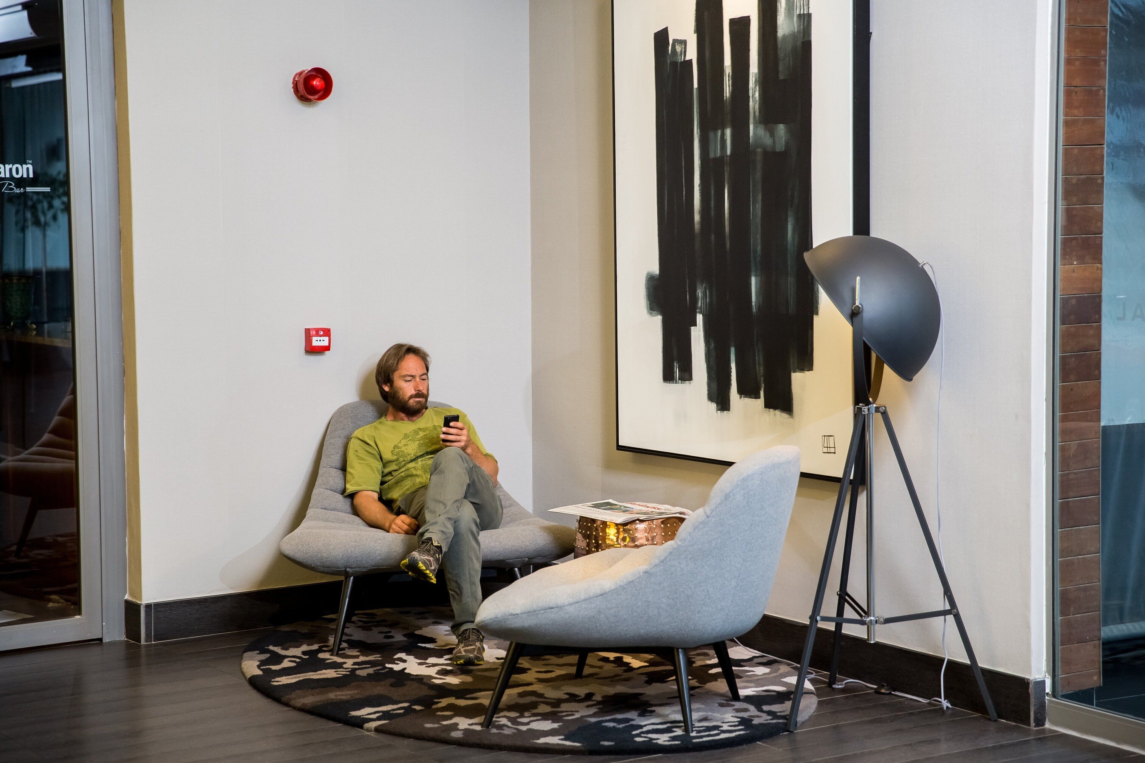 A man sitting on a grey chair in a modern room with a large abstract painting on the wall, a floor lamp, and a small table with a box.