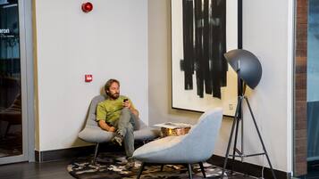 A man sitting on a grey chair in a modern room with a large abstract painting on the wall, a floor lamp, and a small table with a box.