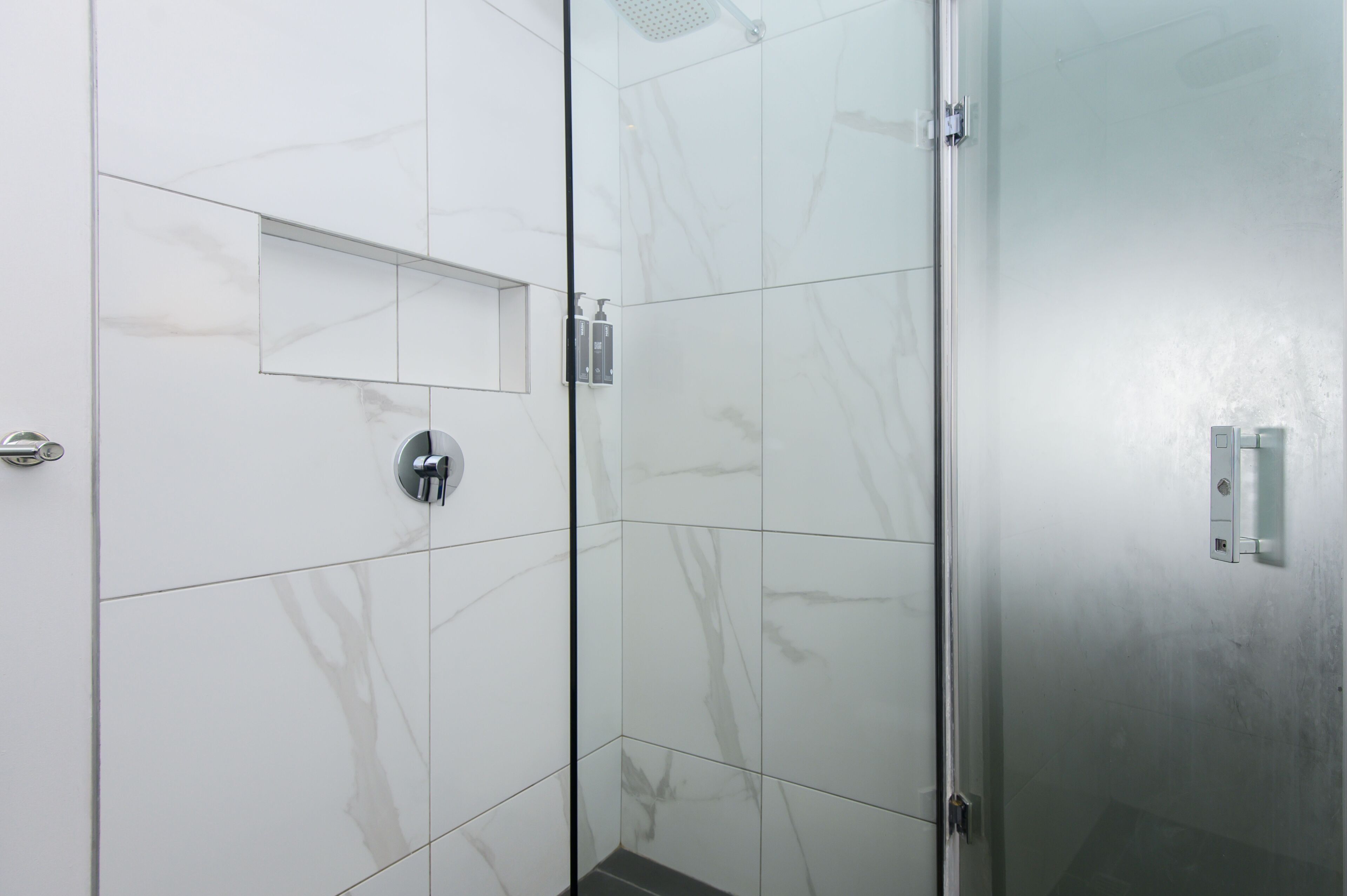 A modern bathroom with white marble tiles, a glass shower enclosure, and a small mirror.