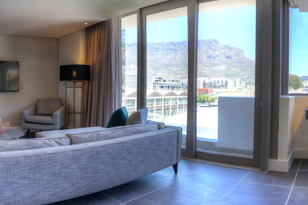 A modern hotel room with a large bed, a chair, a desk, and a view of a cityscape and mountains.