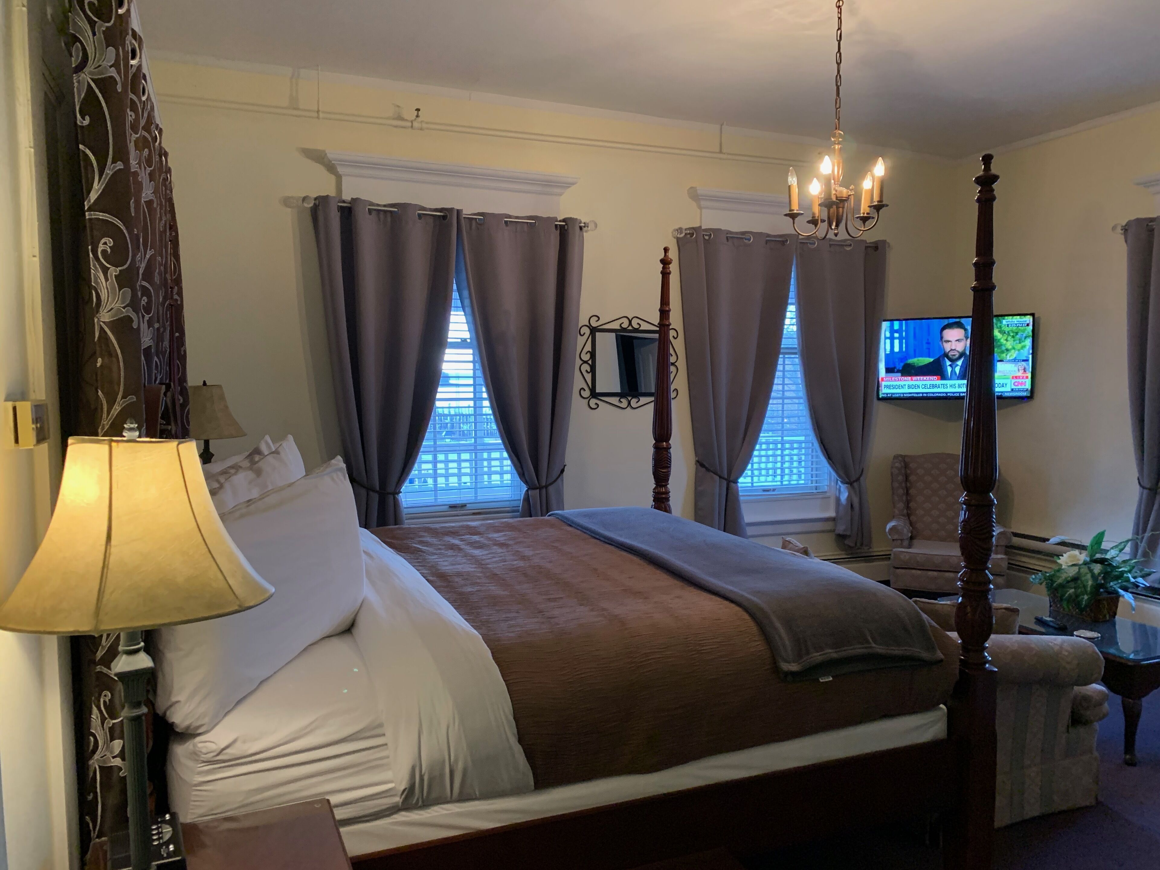 A four-poster bed with a brown bedspread, two bedside lamps, and a television on the wall.