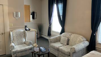 A hotel room with two floral-patterned armchairs, a small round table with a box and a bottle, a bed, a dresser, a mirror, and a window with curtains.