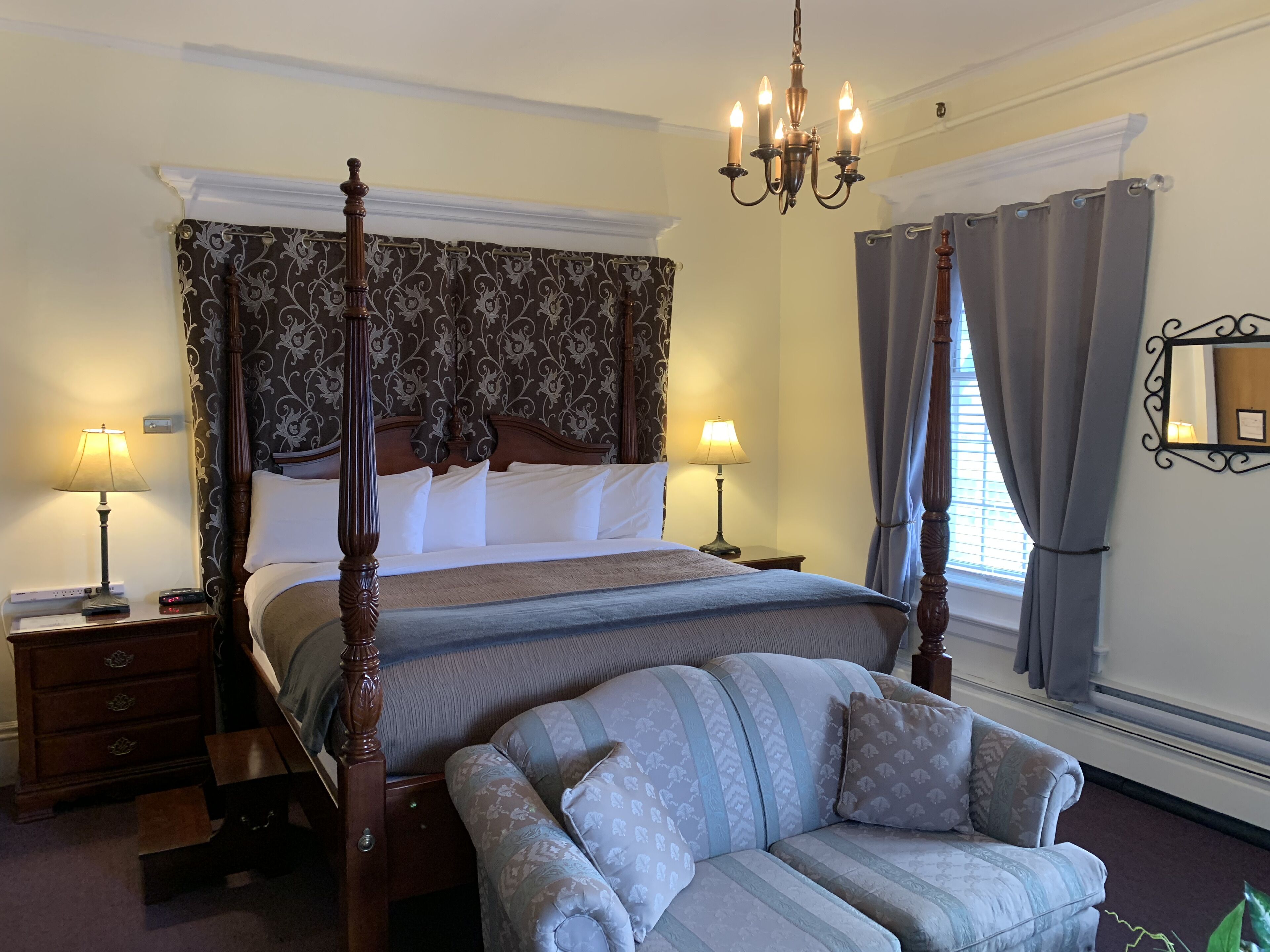 Ein Bett mit Baldachin, ein dunkles, verziertes Kopfteil, zwei Nachttische mit Lampen, ein großes Fenster mit Vorhängen und ein gemustertes Sessel.