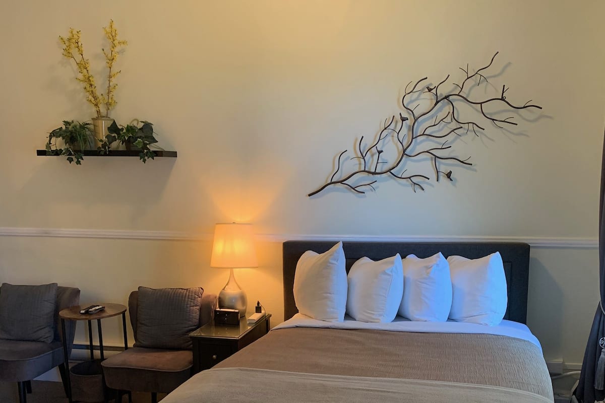 Un dormitorio ordenado con una cama grande, una mesita de noche, una lámpara, una silla y un adorno en la pared.