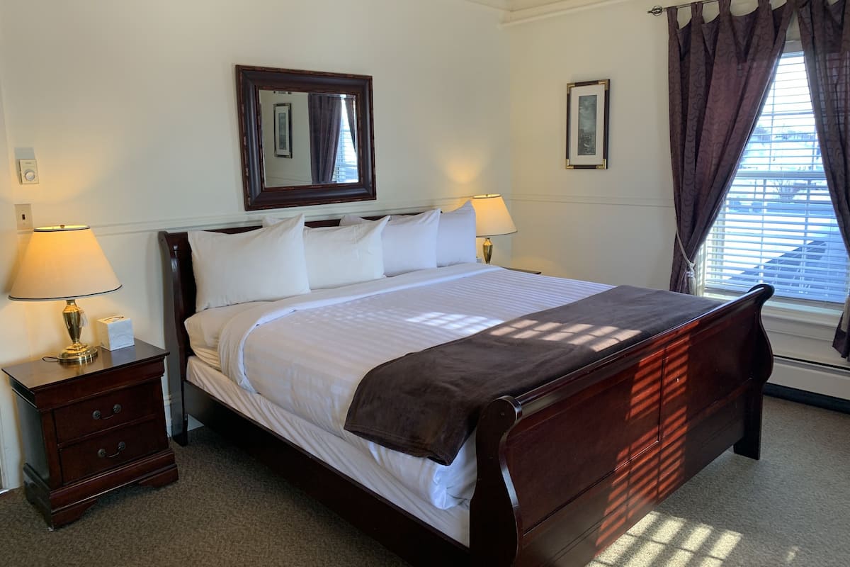 Una cama bien hecha con ropa blanca, dos mesitas de noche con lámparas, un espejo grande sobre la cama y una ventana con cortinas.