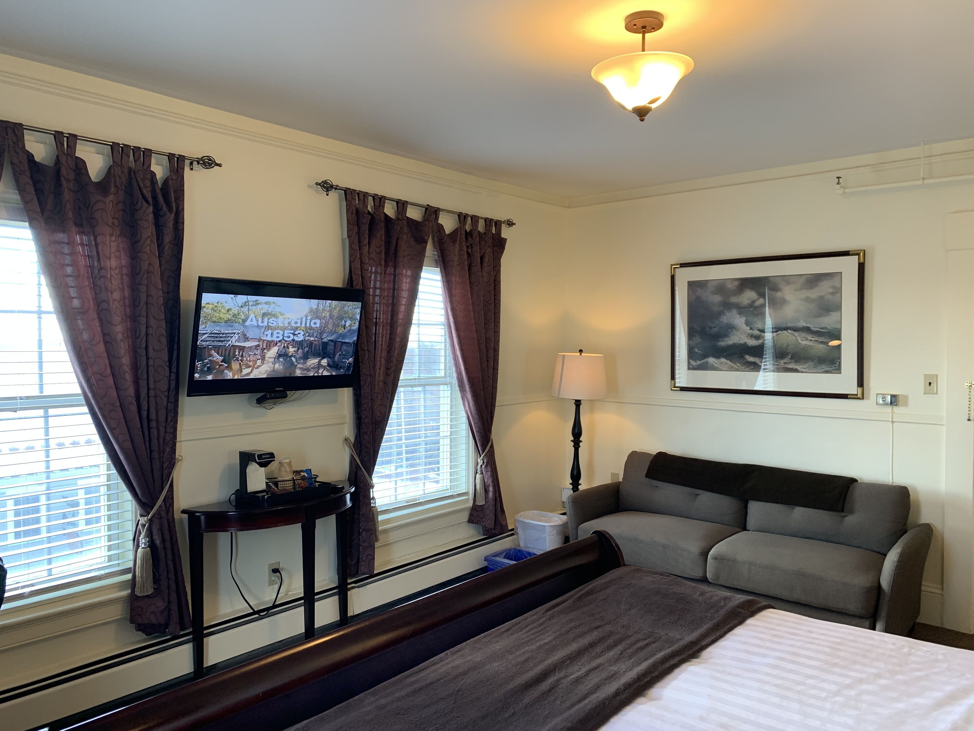 A hotel room with a bed, a television mounted on the wall, a sofa, a lamp, and a window with curtains.