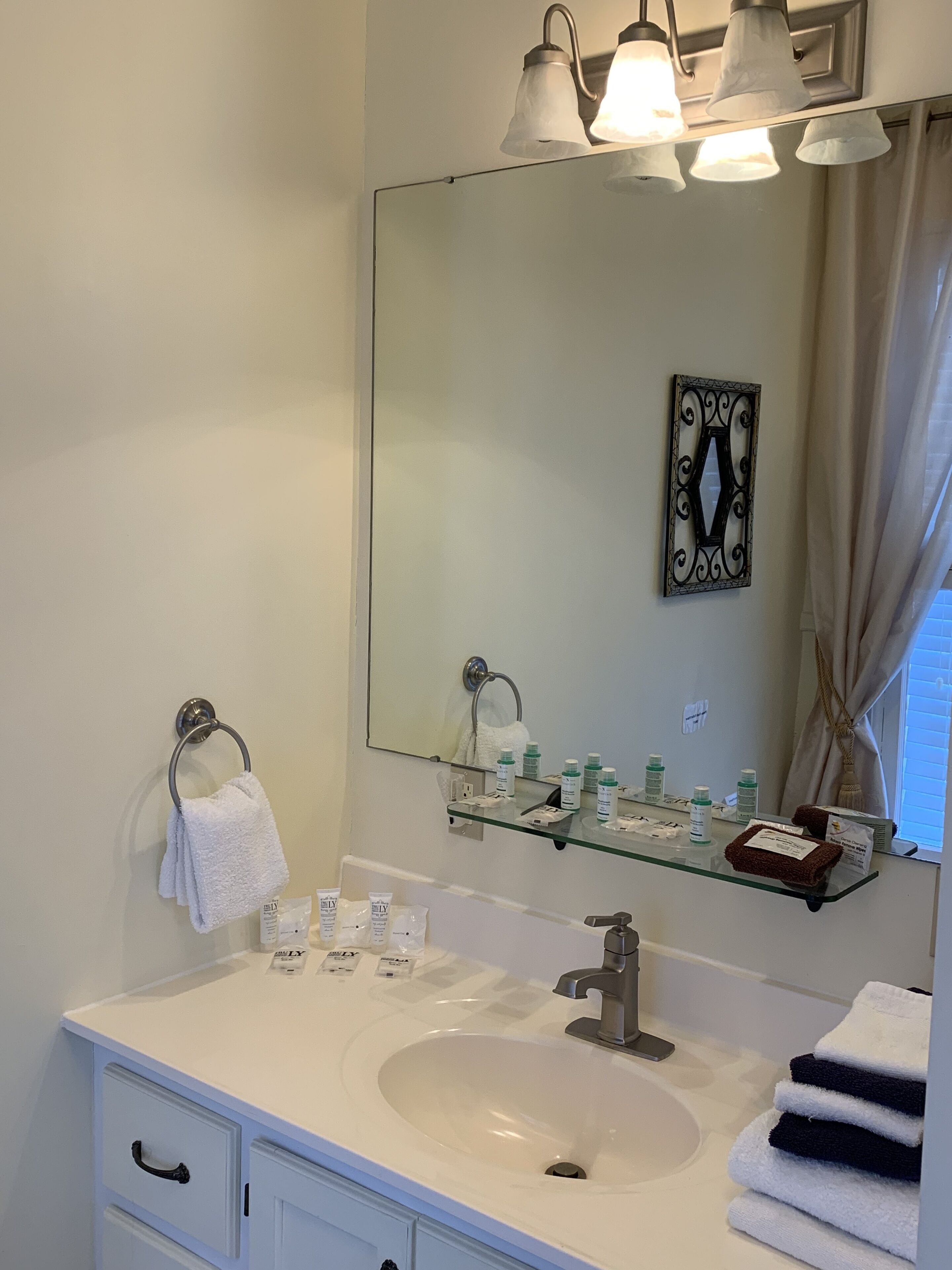 A bathroom with a white sink, a glass shelf with toiletries, a towel ring, and a mirror with a decorative letter frame.